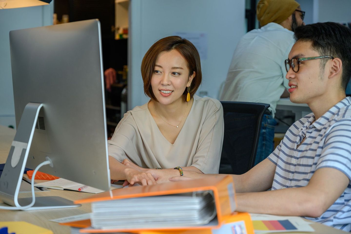 Two colleagues at a desk going through work on a screen beside a stack of binders